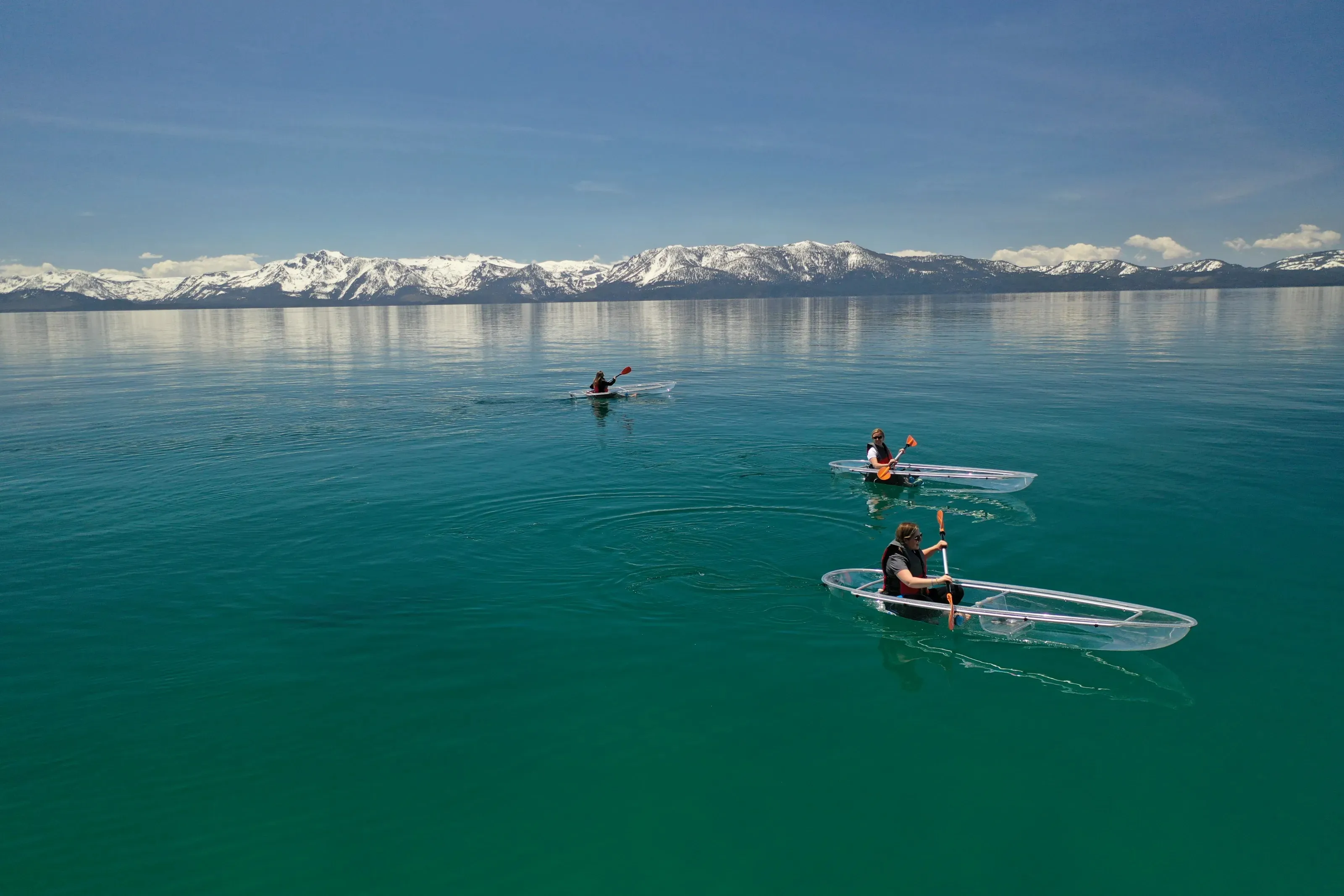 Three clear kayaks crossing calm Lake Tahoe with snow-covered mountains beyond
