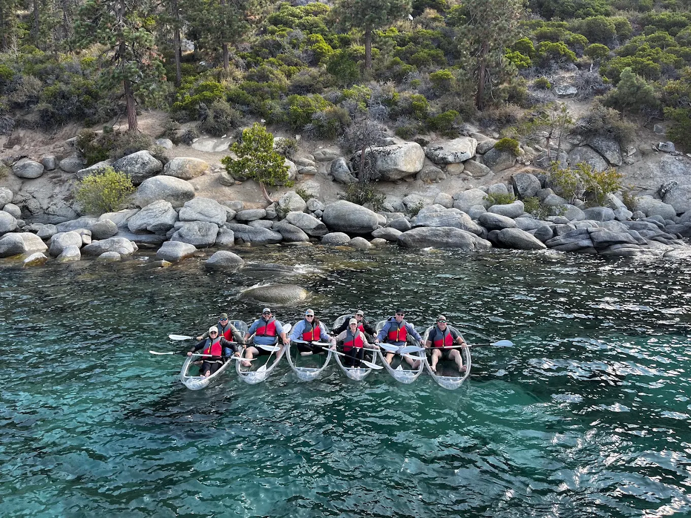 A group of six guests paddling clear kayaks together near the Tahoe shoreline