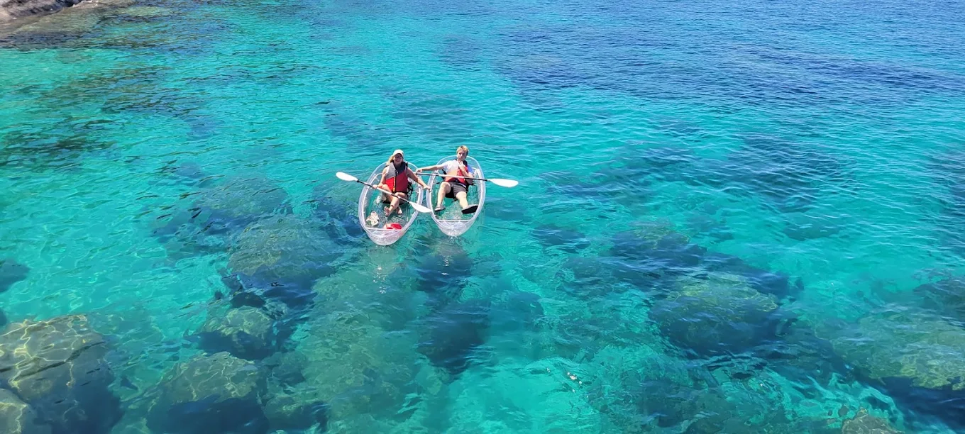 An aerial view of two guests paddling clear kayaks over turquoise water