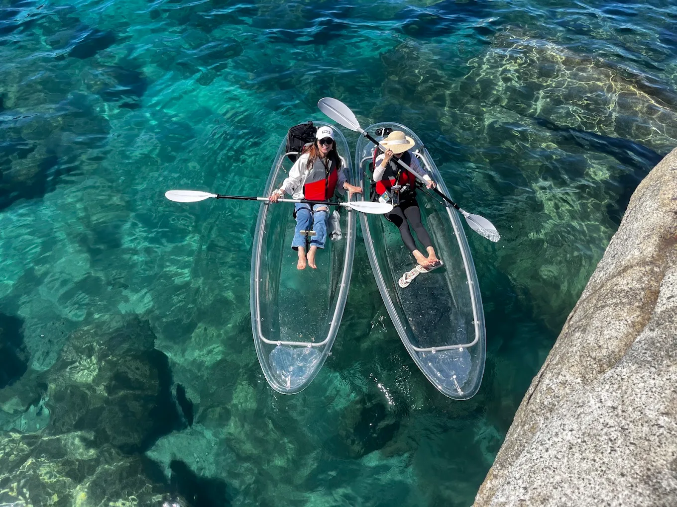 Two guests paddling clear kayaks side by side near a rocky point