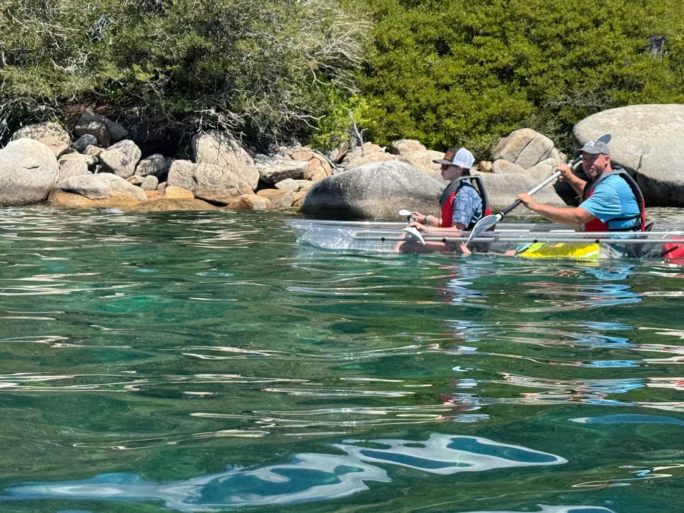 Two guests sharing one fully transparent tandem clear kayak near the rocky Tahoe shoreline